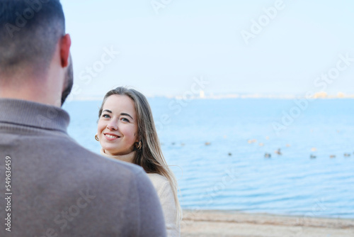 Young man and woman holding hands and posing on the beach against the backdrop of blue water. The girl looks at the guy and smiles.