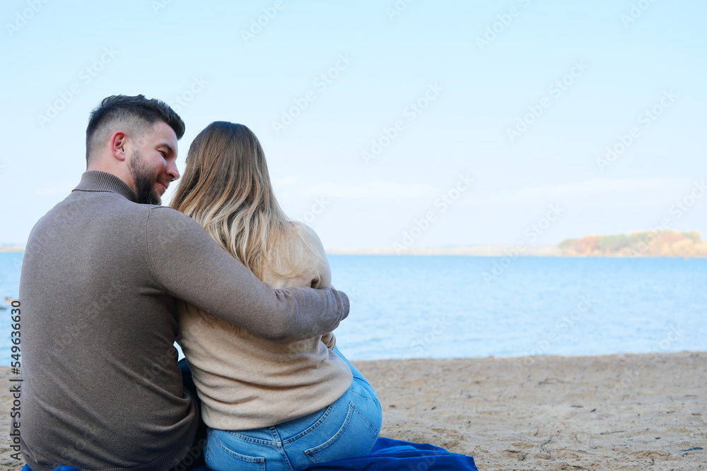 Fototapeta premium A young man and woman are sitting against the backdrop of a large blue lake and hugging. couple sitting with their backs to the camera