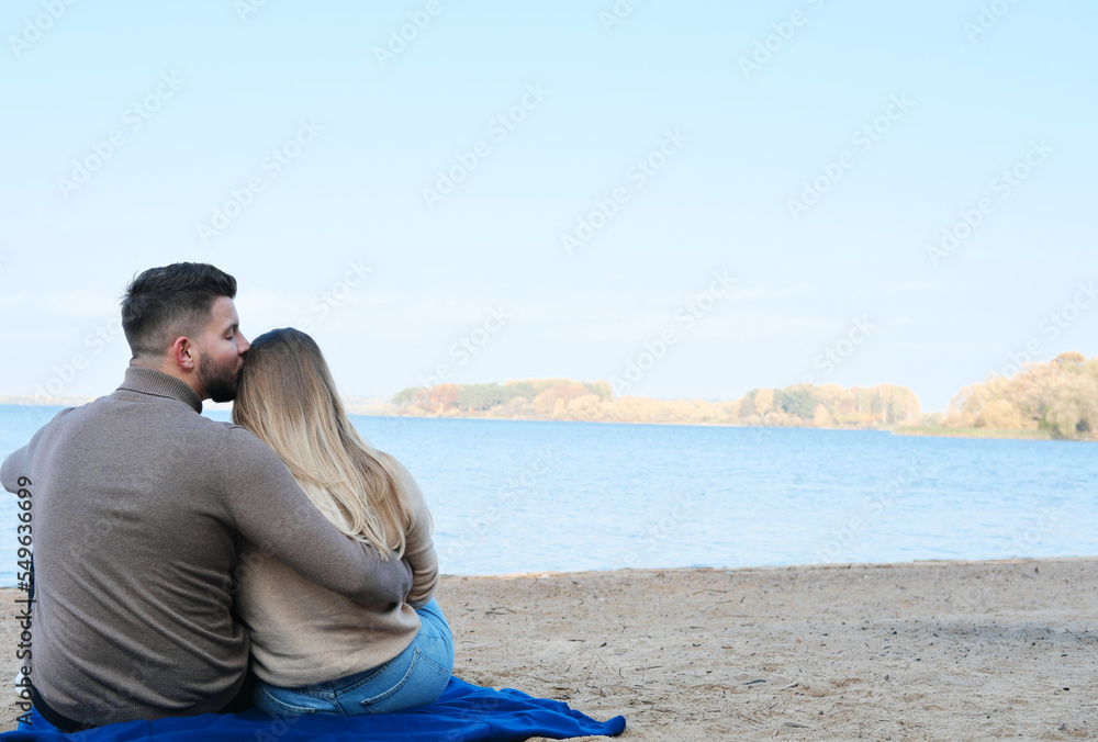 A young man and woman are sitting against the backdrop of a large blue lake and hugging. The couple sits with their backs to the camera, the guy kisses the girl's hair