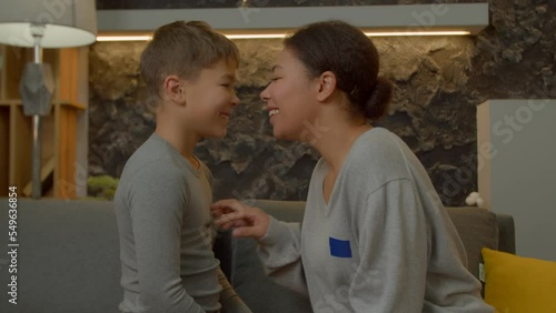 Portrait of carefree affectionate pretty African American mother and joyful multiethnic school age son sitting face to face on couch, having fun rubbing noses, expressing cheerful mood and happiness.