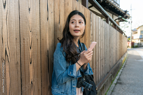 asian Japanese girl tourist using online map on phone while taking break against wooden wall in a neighborhood in uji Kyoto japan. she looks into distance and thinks about her next destination.
