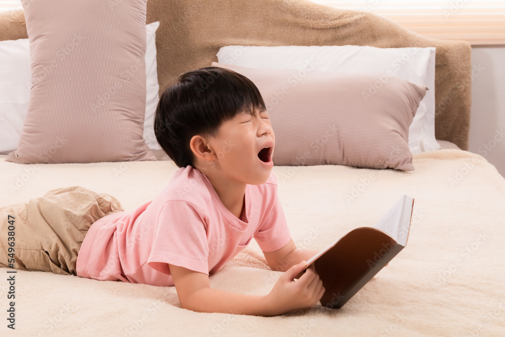 Asian little child boy feeling sleepy yawn and rubbing both eyes with both hands while reading favorite manga, elementary children tired after school lying on bed and read book, doing homework. Asian little child boy feeling sleepy yawn and rubbing both eyes with both hands while reading favorite manga, elementary children tired after school lying on bed and read book, doing homework.