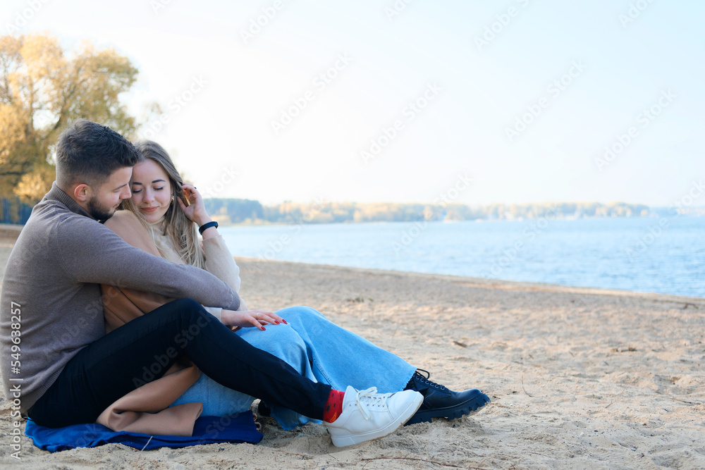 Date on the beach in the cold season. The guy hugs the girl from behind by the shoulders and tries to warm her
