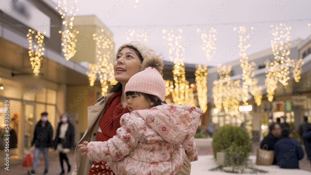 mother holding her baby daughter is feeling joy while turning around with hand out under glowing Christmas hanging lights outside a shopping mall
