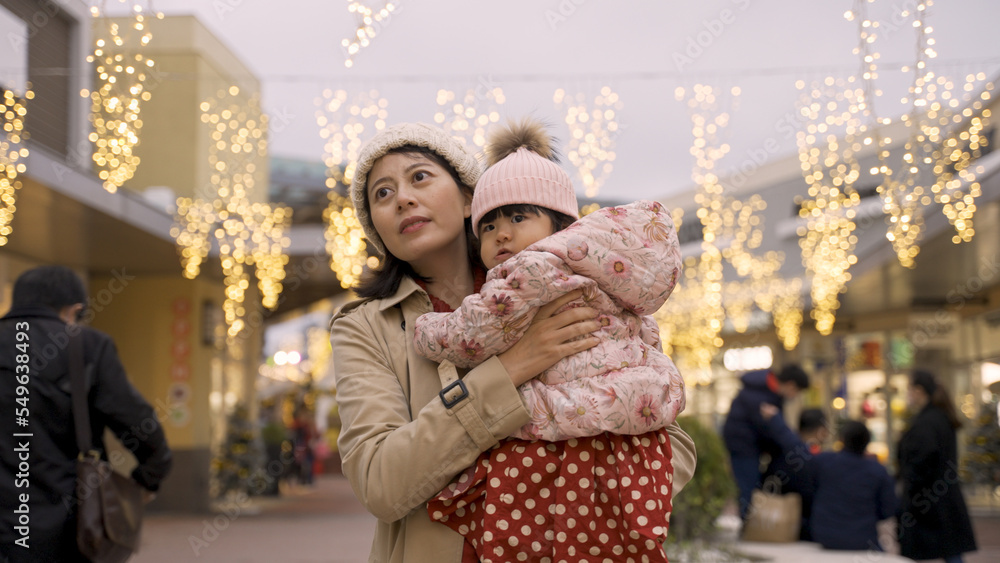 asian mother turning around with baby in arms while having joy moments against background of a glowing pedestrian shopping area with lights in Christmas season