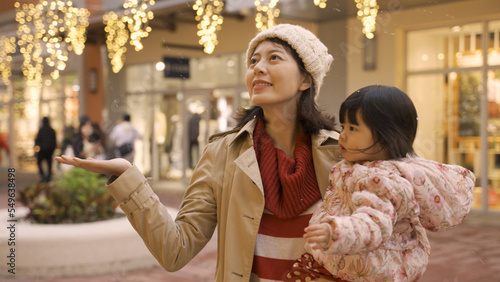 asian mother holding excited baby daughter with her hand out to catch the snow in air while they are doing Christmas shopping on city street with festive light