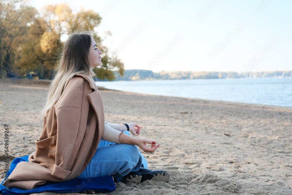 A young woman in a brown coat sits in a lotus position on the beach. The concept of finding a state of inner peace and happiness.