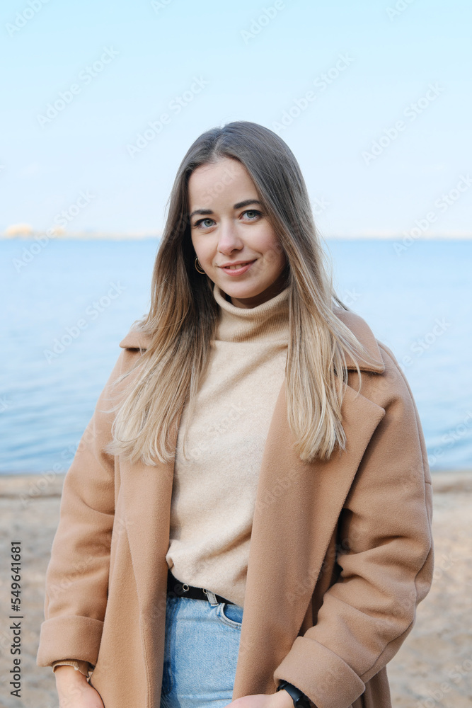 Beautiful young woman in a brown coat posing on the beach. Girl smiling and looking at the camera Beautiful young woman in a brown coat posing on the beach. Girl smiling and looking at the camera