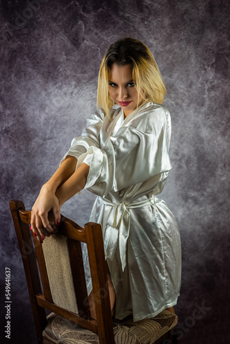 Beautiful adult sexy woman sitting on a chair in a white silk robe posing in the studio.