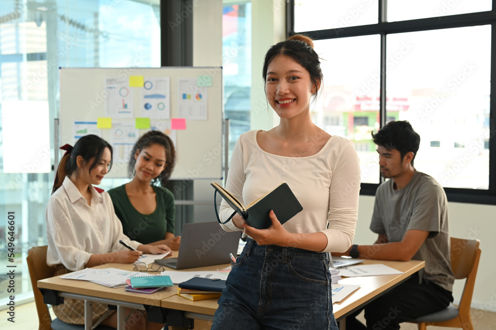 Fototapeta premium Positive young female team leader standing in front of meeting table at creative office and smiling to camera