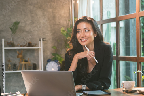 Asian female employee working on laptop plan to present for her client company, young businesswoman sitting happy and smiling at office thinking new product with modern idea.