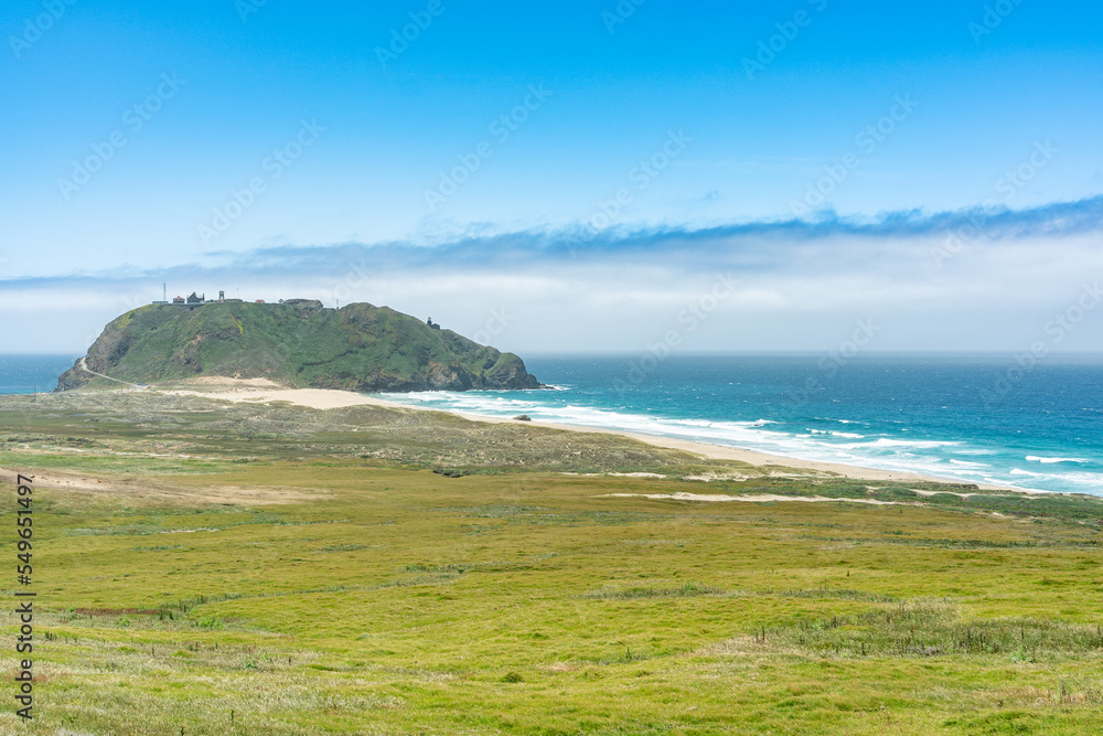 California, USA - May 19, 2018: grass covered land meeting the sea and a distant island on the pacific ocean under blue sky in big sur