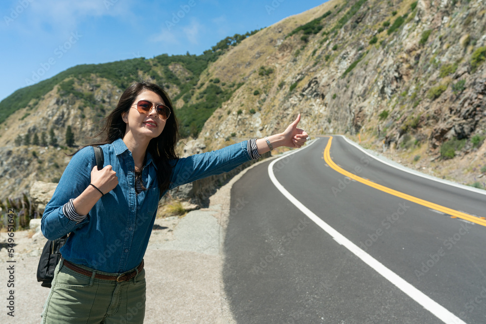 cheerful asian Korean girl hitchhiker sticking thumb out for free ride at roadside on hillside highway