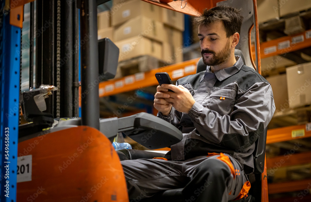 Young wearhouse forklift worker, having small break from work and using phone while sitting on forklift