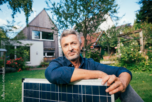 Smiling mature man sitting with solar panel in front of house