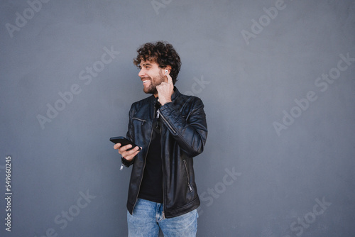 Happy man holding mobile phone adjusting wireless in-ear headphones in front of gray wall
