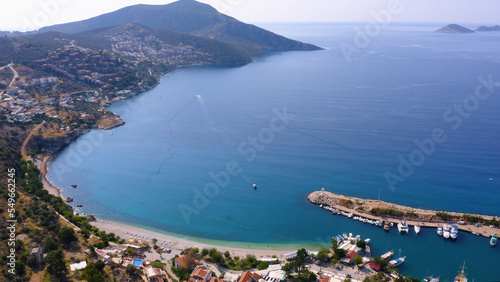 Aerial drone view of coastal town with green mountains. Turquoise sea bay with boats anchored at harbor. Antalya province, Turkey.