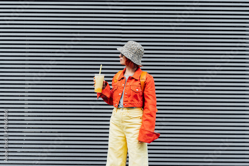 Stylish girl in bright clothes drinking sugar flavored tapioca bubble tea while walking near gray striped urban wall. Full length portrait of fashionable hipster girl. Street fashion. Copy space © okrasiuk