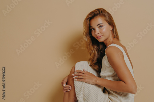 Glamorous young tanned blonde woman sitting sideways with legs up on top of chair in studio photo