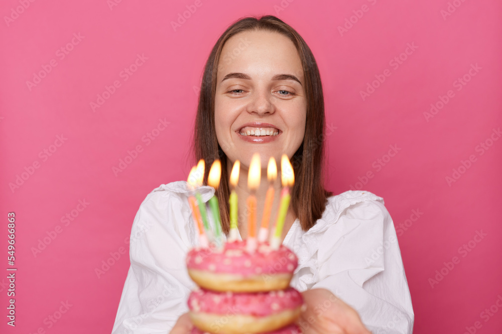 Portrait of cute charming woman with brown hair wearing white blouse holding donut, blowing candles, looking at dessert with toothy smile, enjoying her birthday, posing isolated over pink background. Portrait of cute charming woman with brown hair wearing white blouse holding donut, blowing candles, looking at dessert with toothy smile, enjoying her birthday, posing isolated over pink background.