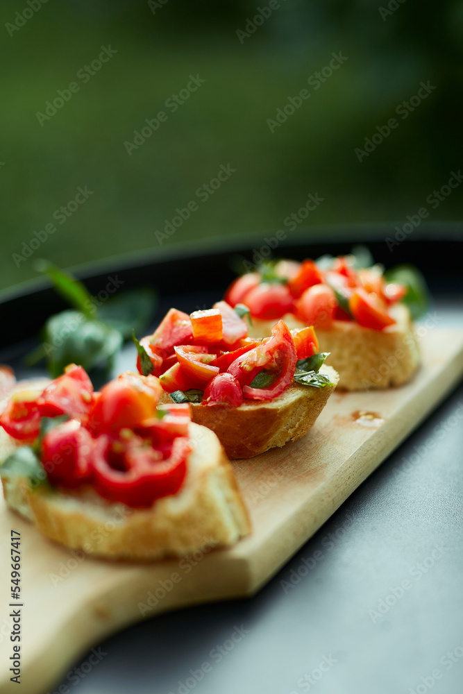 Italian bruschetta with fresh red tomatoes and basil herbs on a wooden board outdoor on the yard, backyard at home, sunlight. Classic Italy food, antipasto Italian bruschetta with fresh red tomatoes and basil herbs on a wooden board outdoor on the yard, backyard at home, sunlight. Classic Italy food, antipasto