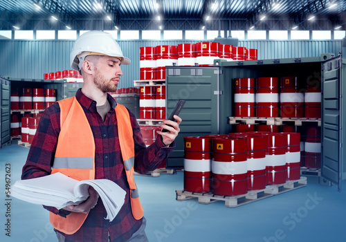 Man warehouse agent. Manager with phone and documentation. Barrels of oil on wooden pallets. Guy works in warehouse with oil. Containers with barrels behind man. Supervisor in industrial hangar