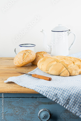 View of rustic bread loaf and rolls on cloth and wooden table with knife, coffee pot and cup, white background, vertical, with copy space