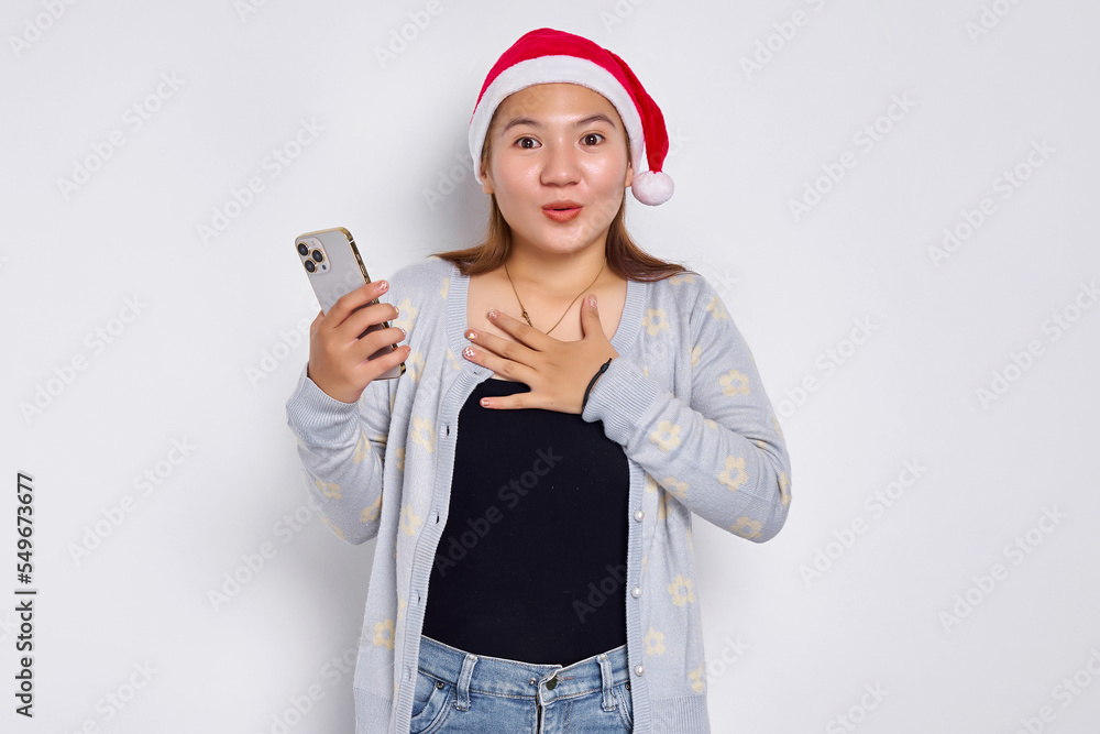 Excited young Asian woman in a Christmas hat holding mobile phone and holding chest isolated on white background Excited young Asian woman in a Christmas hat holding mobile phone and holding chest isolated on white background