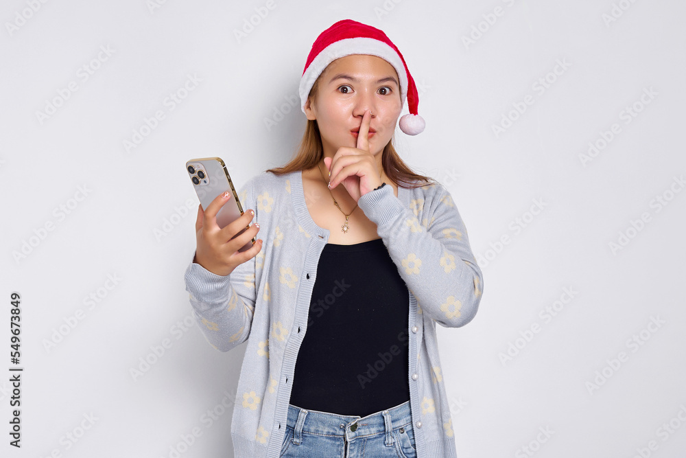 Serious young Asian woman in a Christmas hat holding mobile phone, making silence gesture isolated on white background Serious young Asian woman in a Christmas hat holding mobile phone, making silence gesture isolated on white background