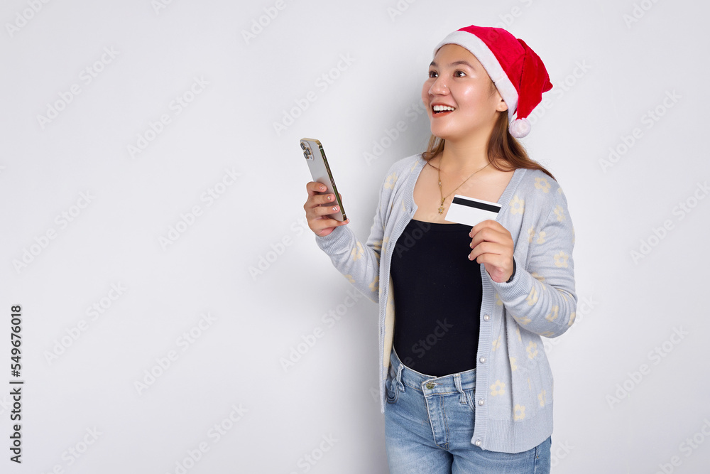 Cheerful young Asian woman in a Christmas hat holding mobile phone and blank white credit card isolated on white background Cheerful young Asian woman in a Christmas hat holding mobile phone and blank white credit card isolated on white background