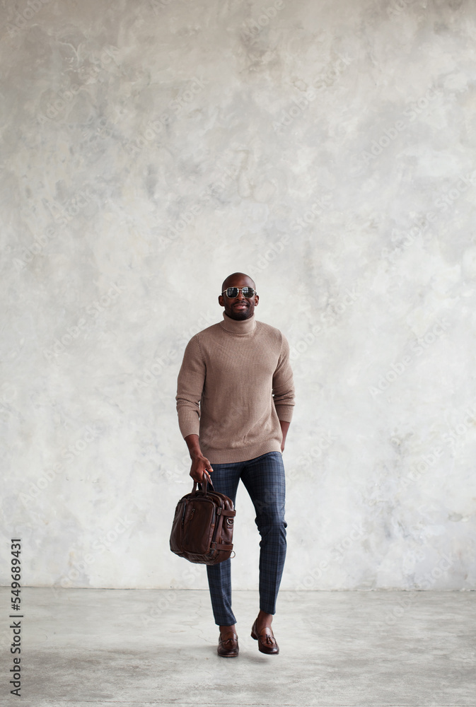 Fashionable full-length man walks against gray wall, wears stylish clothes, brown leather bag and loafers, turtleneck sweater. Men Fashion style