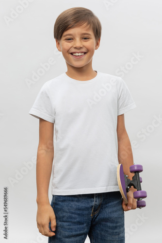 Obraz na płótnie Cool little boy carrying a skateboard and looking at the camera isolated on white background w salonie