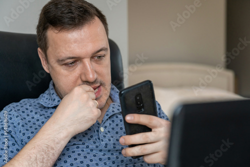 Thoughtful freelancer looking at mobile phone while sitting at desk in hotel. Mature nervous man sitting and working on laptop, bitting his nails and looking at screen of smartphone.