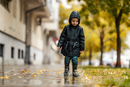 A little boy is splashing rain puddle on the city street during autumn season.