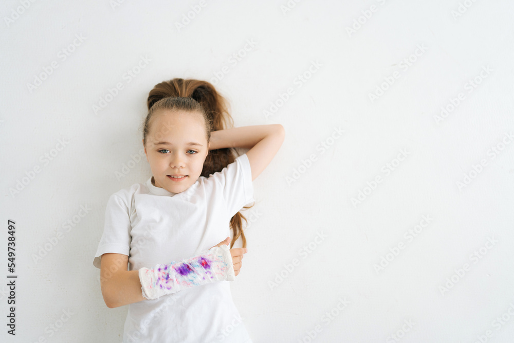High-angle view of positive sweet little girl with broken hand wrapped in plaster bandage with colorful draw lying on white floor in studio looking at camera. Concept of child insurance and healthcare High-angle view of positive sweet little girl with broken hand wrapped in plaster bandage with colorful draw lying on white floor in studio looking at camera. Concept of child insurance and healthcare