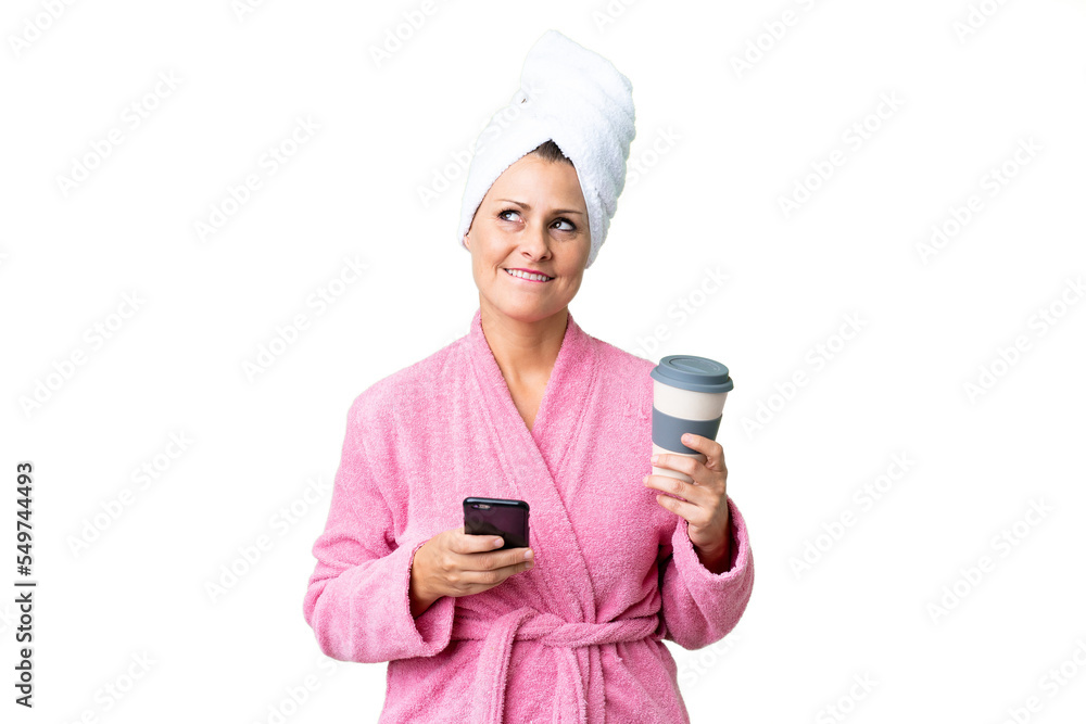 Middle age caucasian woman in a bathrobe over isolated background holding coffee to take away and a mobile while thinking something Middle age caucasian woman in a bathrobe over isolated background holding coffee to take away and a mobile while thinking something