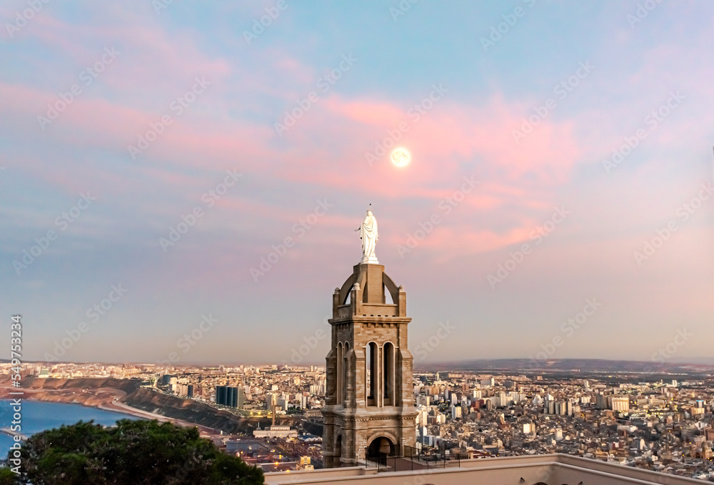 Aerial view from Santa Cruz Chapel tower and the virgin Mary statue back from behind looking at a full moon and the city of Oran with a colorful cloudy sky blue hour sunset twilight and water seaport
