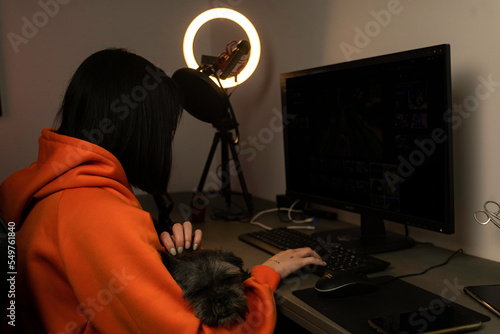 Young gamer girl accompanied by her little long-haired dog is using her computer. She is wearing an orange sweatshirt and is in her gamer room while using her light ring. photo