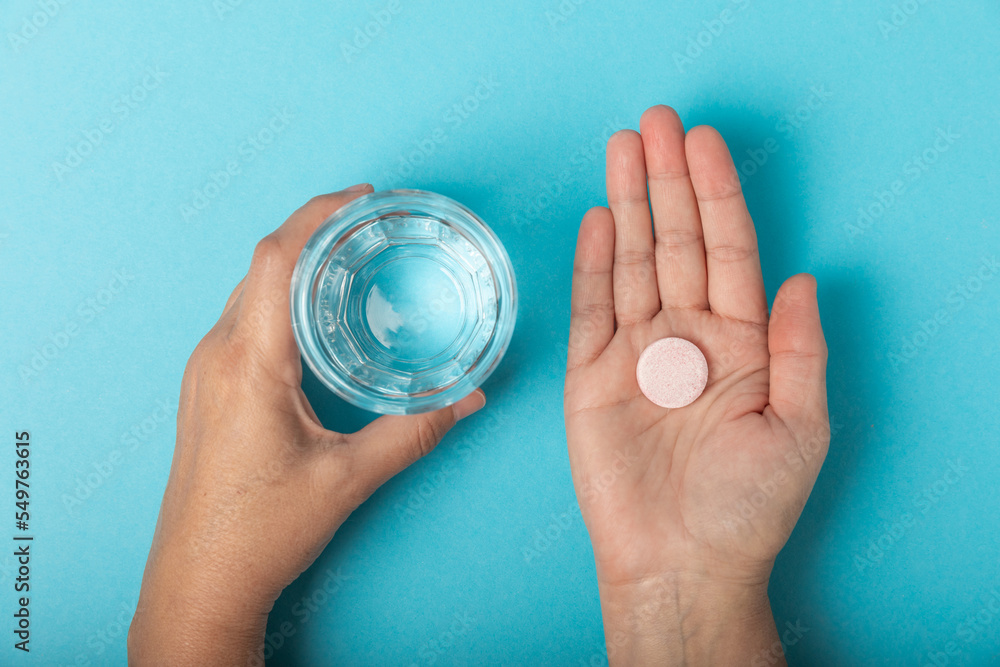 Fototapeta premium Woman's hands hold vitamin pill and glass of water on blue background.Ready to take medicine for headache and cold, painkillers, nutritional supplements,immunity pill.Health care concept.