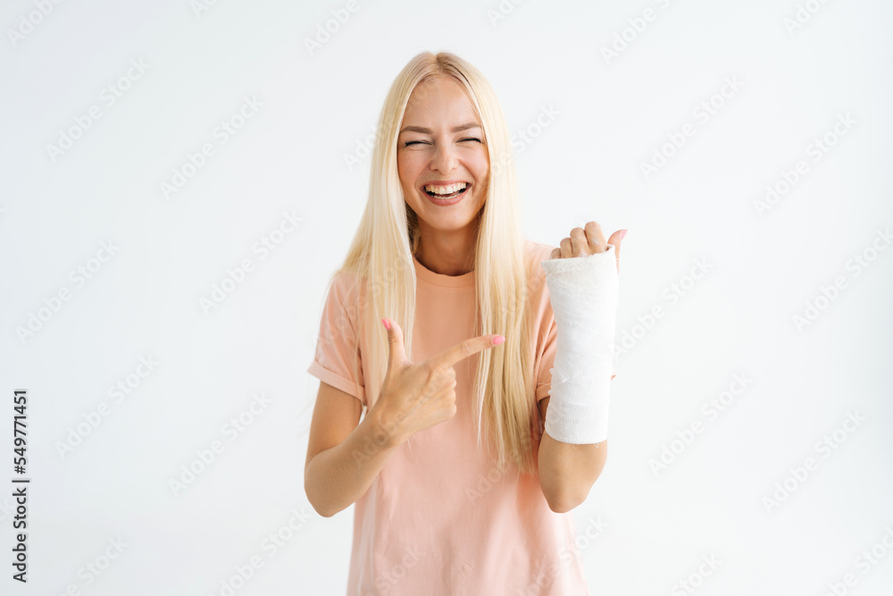 Obraz premium Laughing blonde young woman pointing index fingers to broken arm wrapped in plaster bandage looking at camera, standing on white isolated background. Concept of insurance and healthcare.