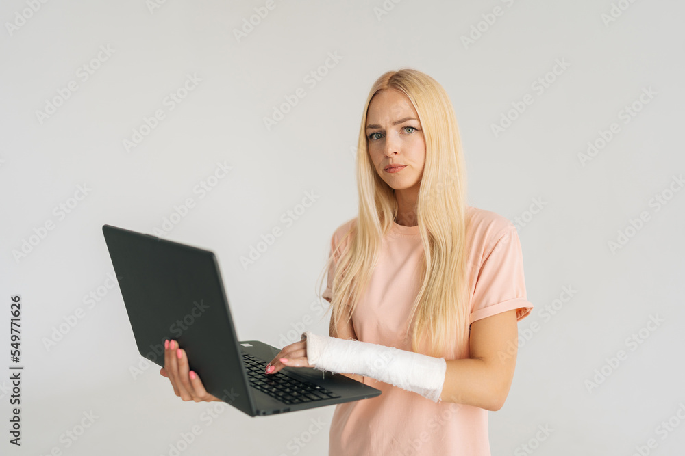 Portrait of serious blonde young woman with broken arm wrapped in plaster bandage holding laptop, looking at camera, standing on white isolated background. Concept of insurance and healthcare Portrait of serious blonde young woman with broken arm wrapped in plaster bandage holding laptop, looking at camera, standing on white isolated background. Concept of insurance and healthcare