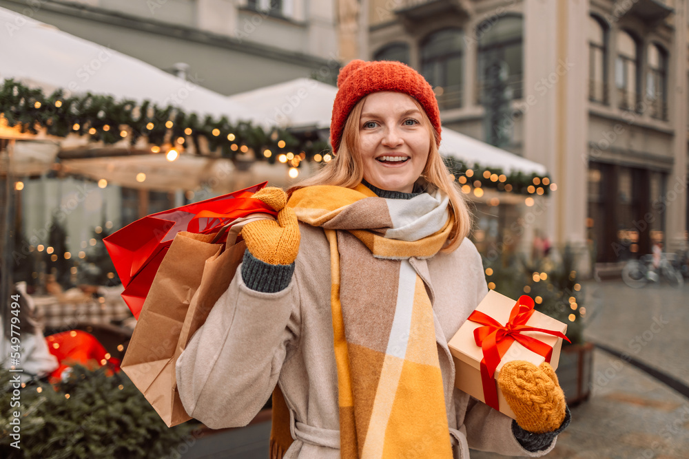 Beautiful happy blonde girl hold shopping bags and gift boxes in hands and smile on city street. Young attractive cheerful woman with paper bags and present box after Christmas shopping in mall