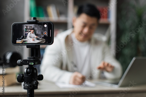 Focus on smartphone screen, smiling asian man talking and gesturing in front of modern smartphone camera, writing report while sitting on background of modern home. Live streaming concept.
