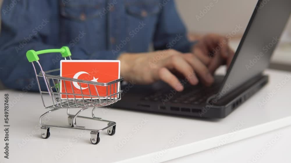 Small shopping cart with flag of Turkey on the table. Man using laptop for shopping online. Small shopping cart with flag of Turkey on the table. Man using laptop for shopping online.