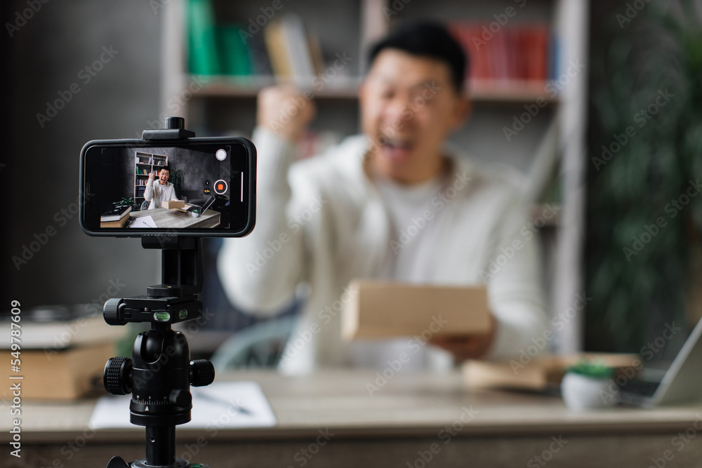 Focus on smart phone screen, blurred young asian man in casual wear recording video using phone while unpacking gift box. Male blogger sharing his emotions with his subscribers in social networks.