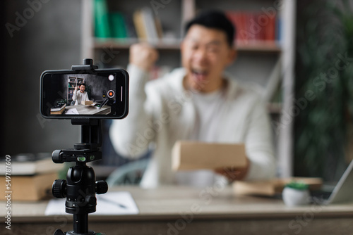 Focus on smart phone screen, blurred young asian man in casual wear recording video using phone while unpacking gift box. Male blogger sharing his emotions with his subscribers in social networks.