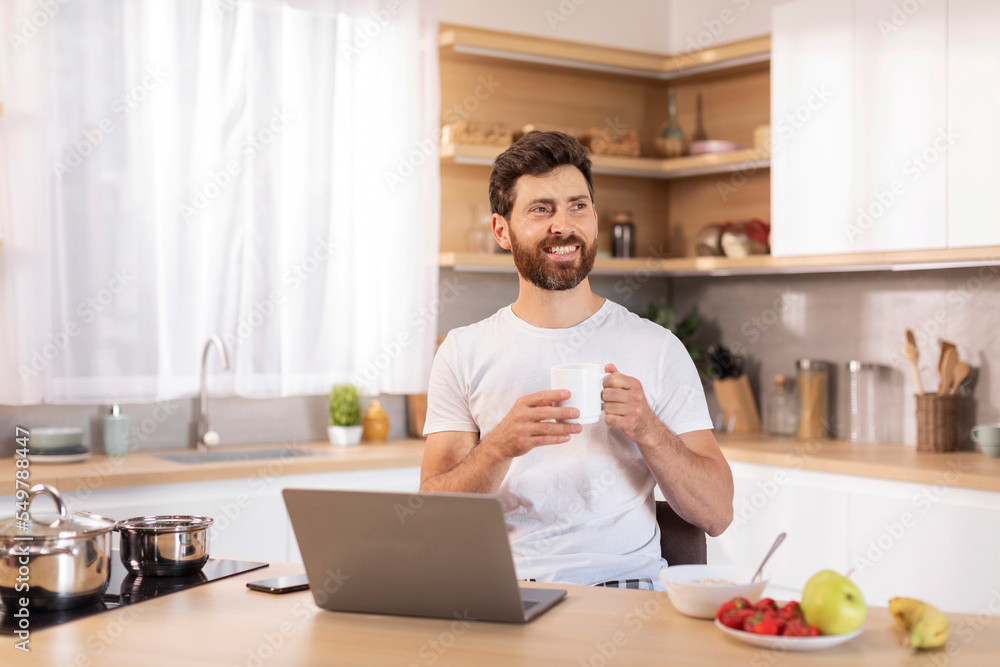 Glad adult caucasian guy with beard in white t-shirt enjoys cup of coffee, sits at table with laptop, thinks Glad adult caucasian guy with beard in white t-shirt enjoys cup of coffee, sits at table with laptop, thinks