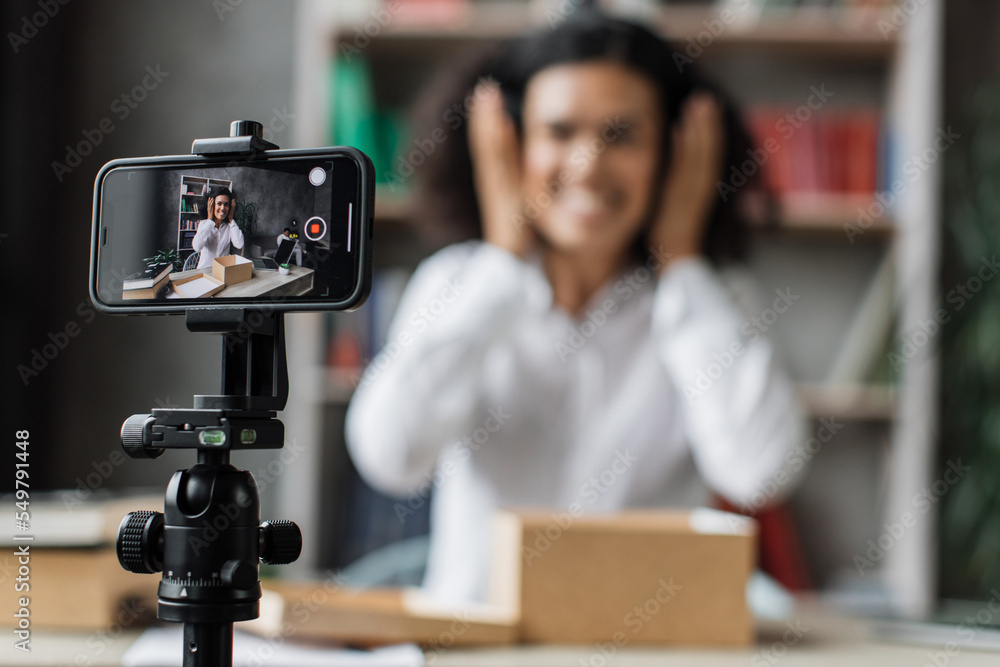 Focus on smart phone screen, woman recording video on phone camera while unpacking box with new wireless headphones. Female influencer sharing with subscribers her positive feedback about new order.