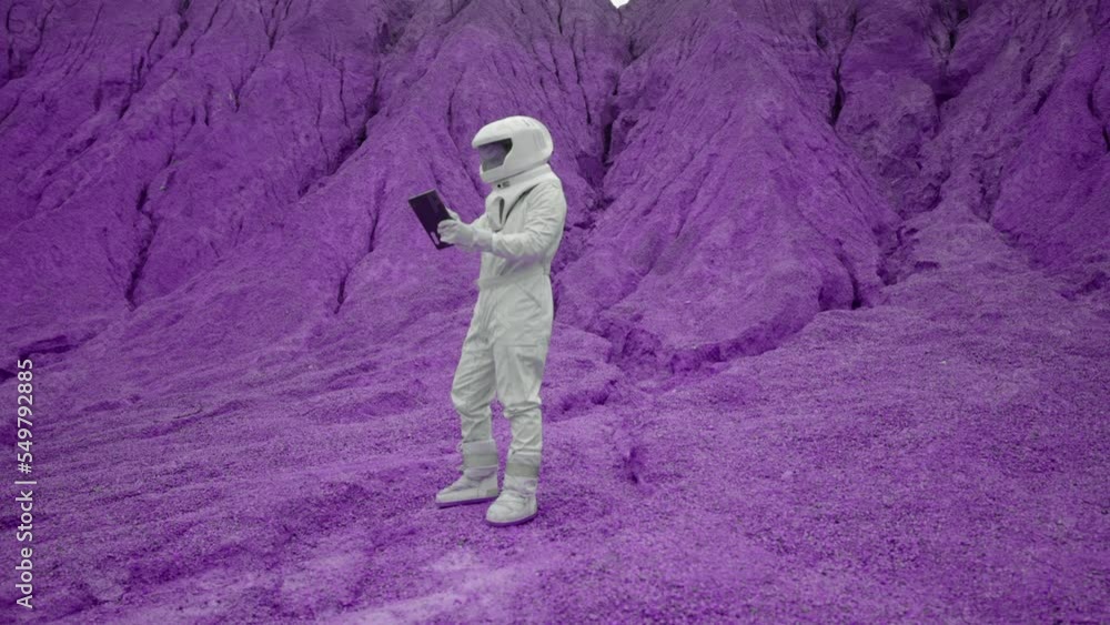 Male astronaut in white suit and helmet working on laptop. Scanning and analysing soil samples for available life on far purple ground planet. Sci-fi futuristic super color concept. Male astronaut in white suit and helmet working on laptop. Scanning and analysing soil samples for available life on far purple ground planet. Sci-fi futuristic super color concept.
