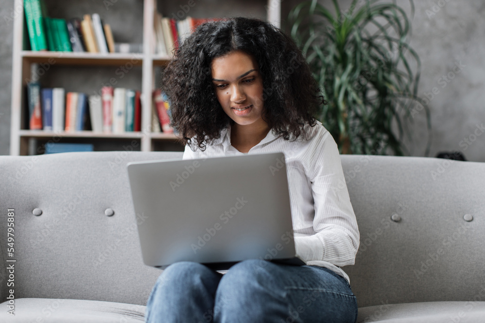 Attractive african american woman in casual clothes sitting on couch and using wireless laptop. Young female browsing internet during leisure time at cozy home. Attractive african american woman in casual clothes sitting on couch and using wireless laptop. Young female browsing internet during leisure time at cozy home.